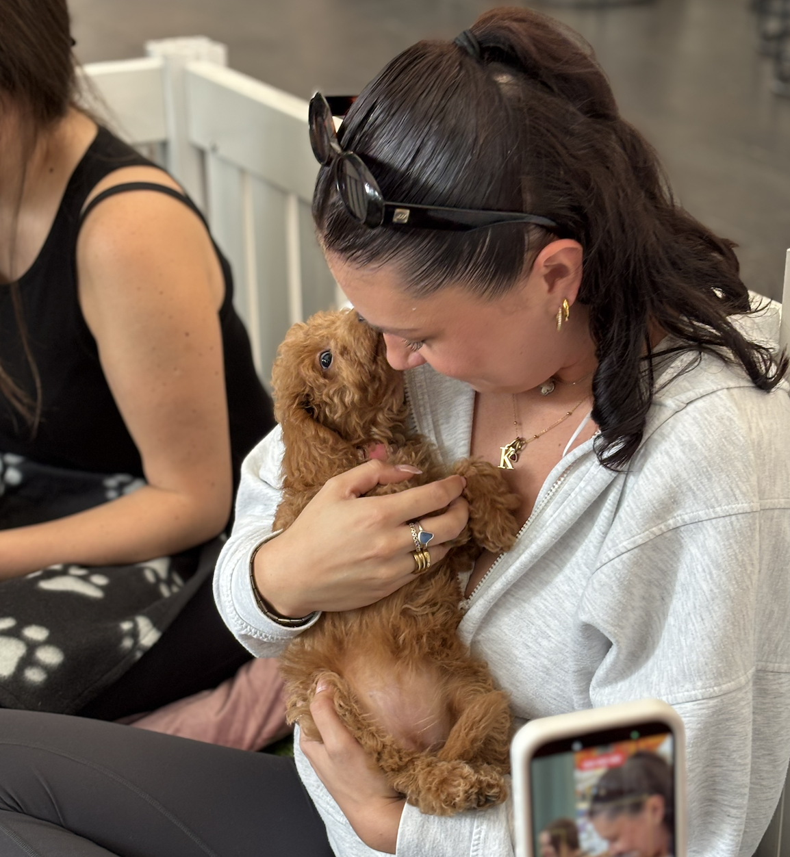A woman holding a small brown poodle puppy close to her face, smiling and looking at it affectionately. The woman has dark hair tied back, sunglasses on her head, and is wearing jewelry. She is seated next to another woman in a black sleeveless top, partially visible. There is a person taking a photo with a smartphone in the foreground.