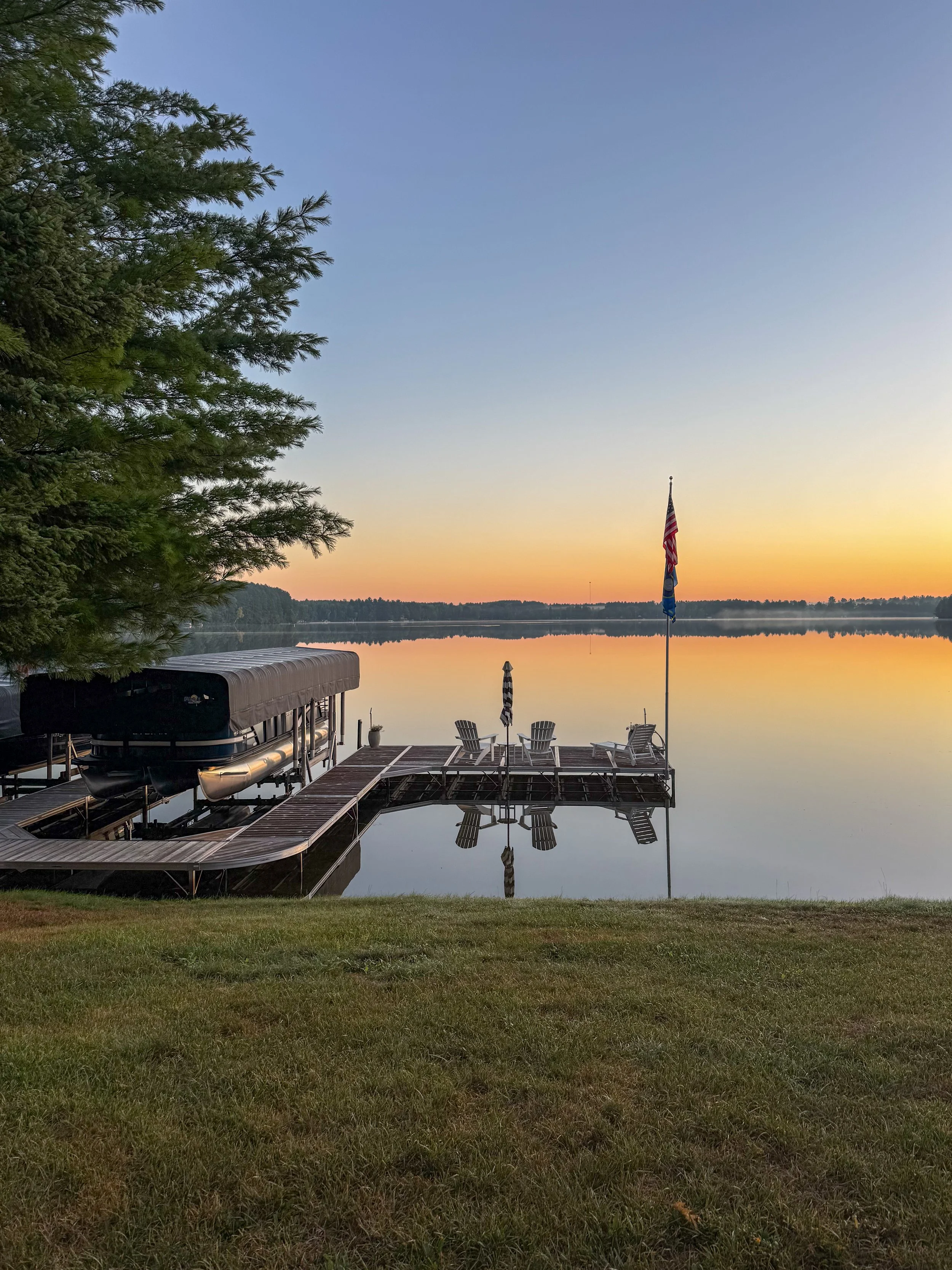 A lakeside scene at sunset with a dock, chairs, and an American flag on a flagpole. The sky is mostly clear with a hint of orange near the horizon, reflecting on the calm water. A large evergreen tree is on the left, partially framing the scene.