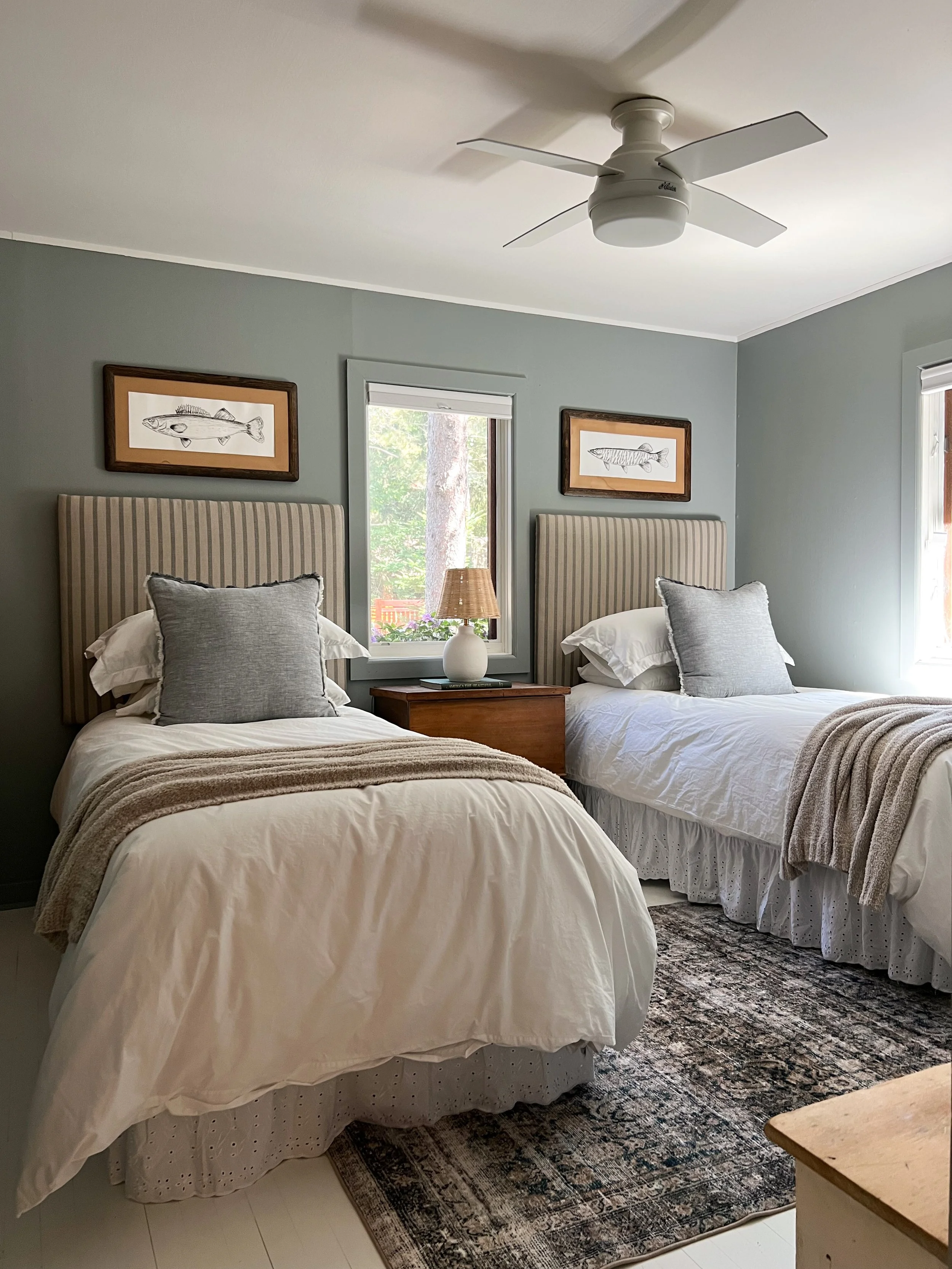 A bedroom with two beds, each with a striped headboard, gray pillows, and white bedding, next to a window and a nightstand with a lamp and a book, decorated with framed fish illustrations and a patterned rug.