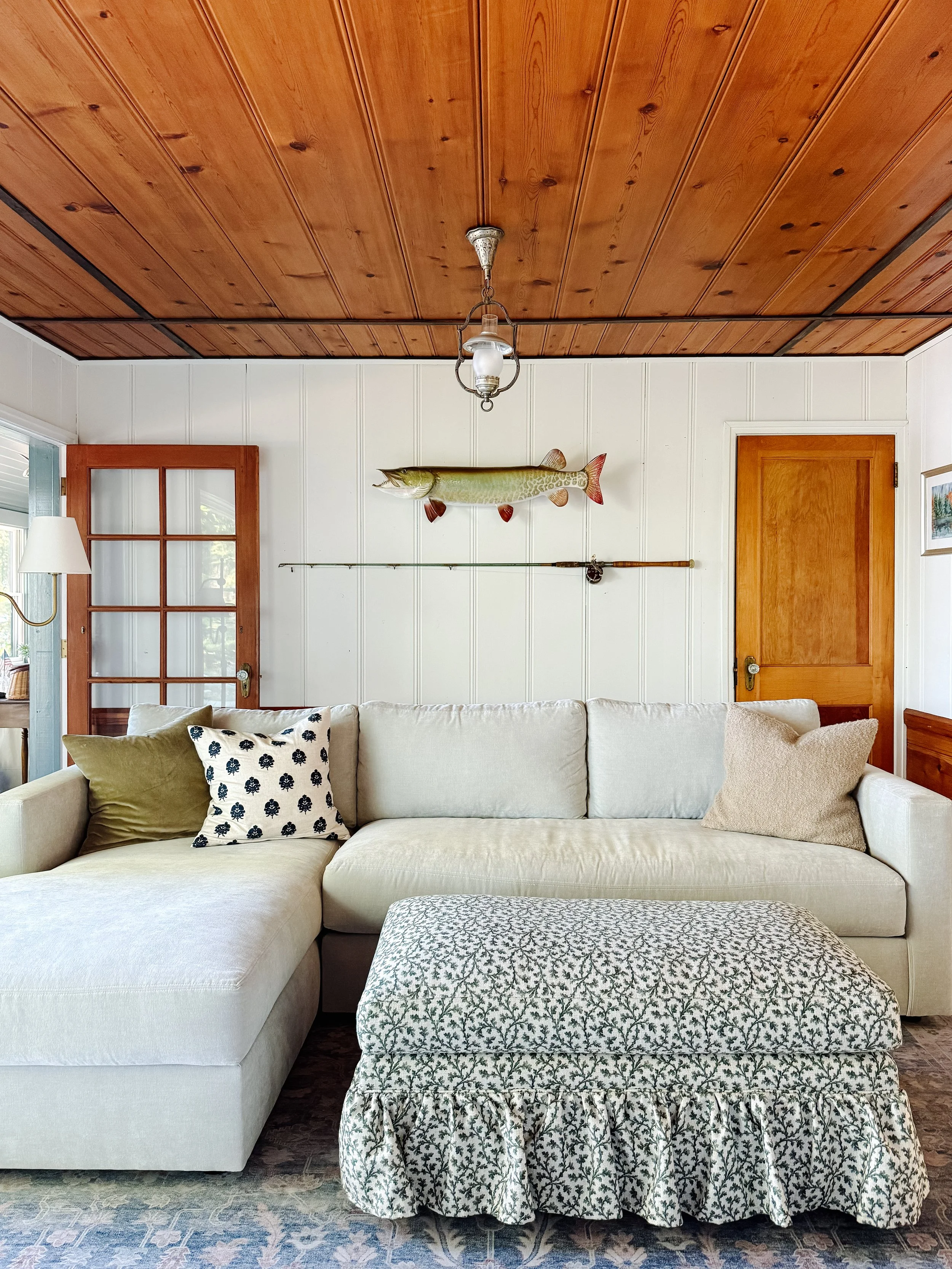 Living room with white sofa, decorative pillows, a patterned ottoman, a wall-mounted painted fish piece, a wooden door, a glass lamp, and a fishing rod on the wall. The ceiling is wood-paneled.