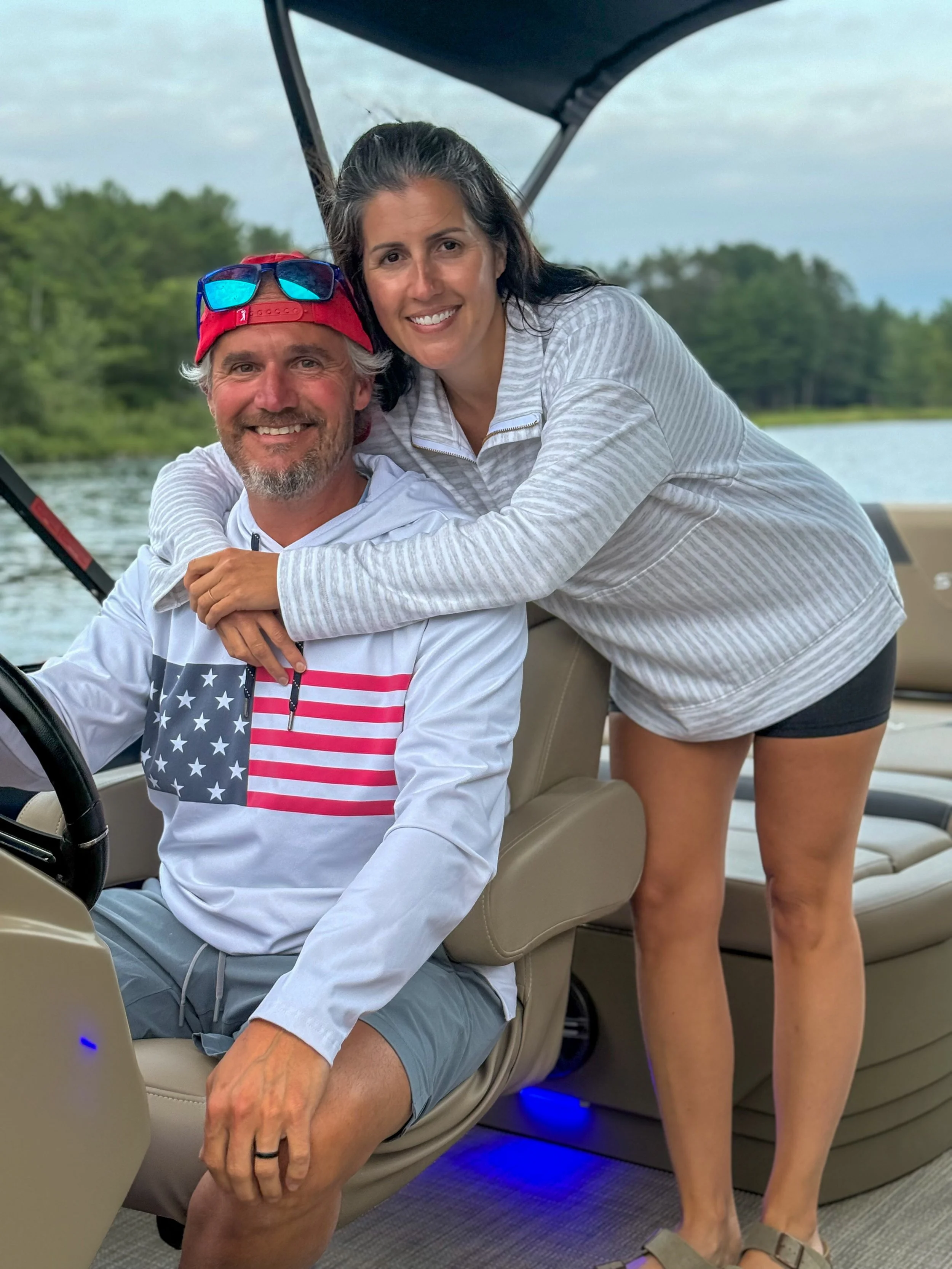 A man sitting on a beige boat seat holding a fishing rod, with sunglasses on his red cap, smiling, and a woman with dark hair smiling and leaning on his shoulder, standing beside him, with a background of water and green trees.