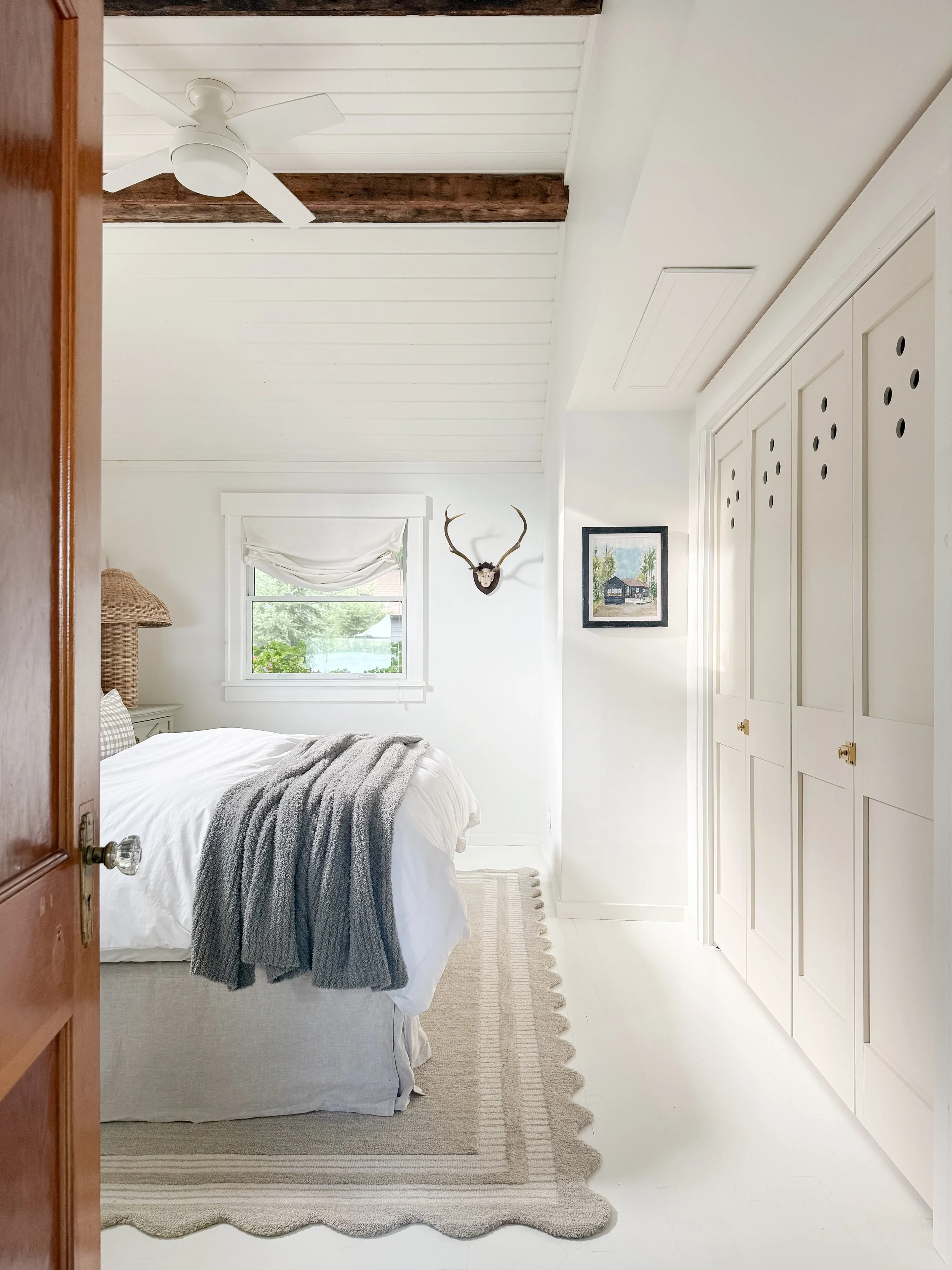 A cozy, well-lit bedroom with a white color scheme, featuring a bed with white bedding and a gray knitted throw, a beige and white striped rug, a window with a white curtain, a painting of a house, a mounted deer antler, and built-in white closet doo