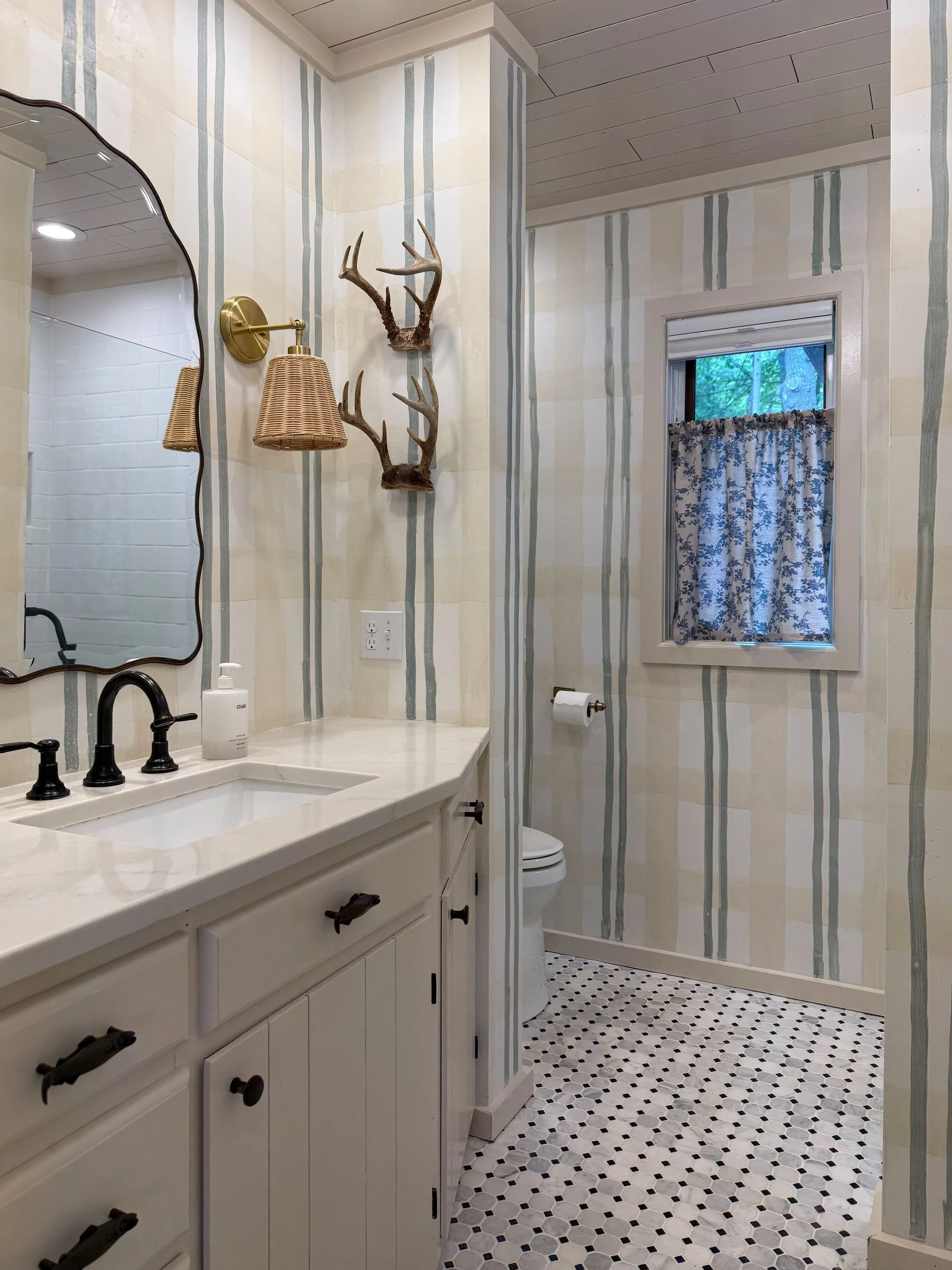 A bathroom with cream-colored walls featuring vertical green and blue stripes. There is a white vanity with black handles, a black faucet, and a soap dispenser. A large mirror with a wavy black frame hangs above the vanity. To the right, there's a wi
