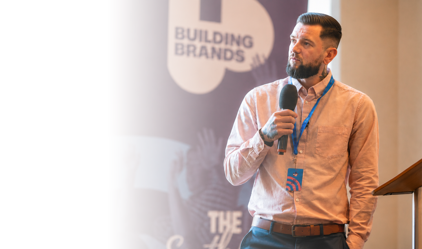 A man with a beard, tattooed hand, and dressed in a pink long-sleeve shirt, holding a microphone, is giving a presentation at a conference called 'Building Brands'.