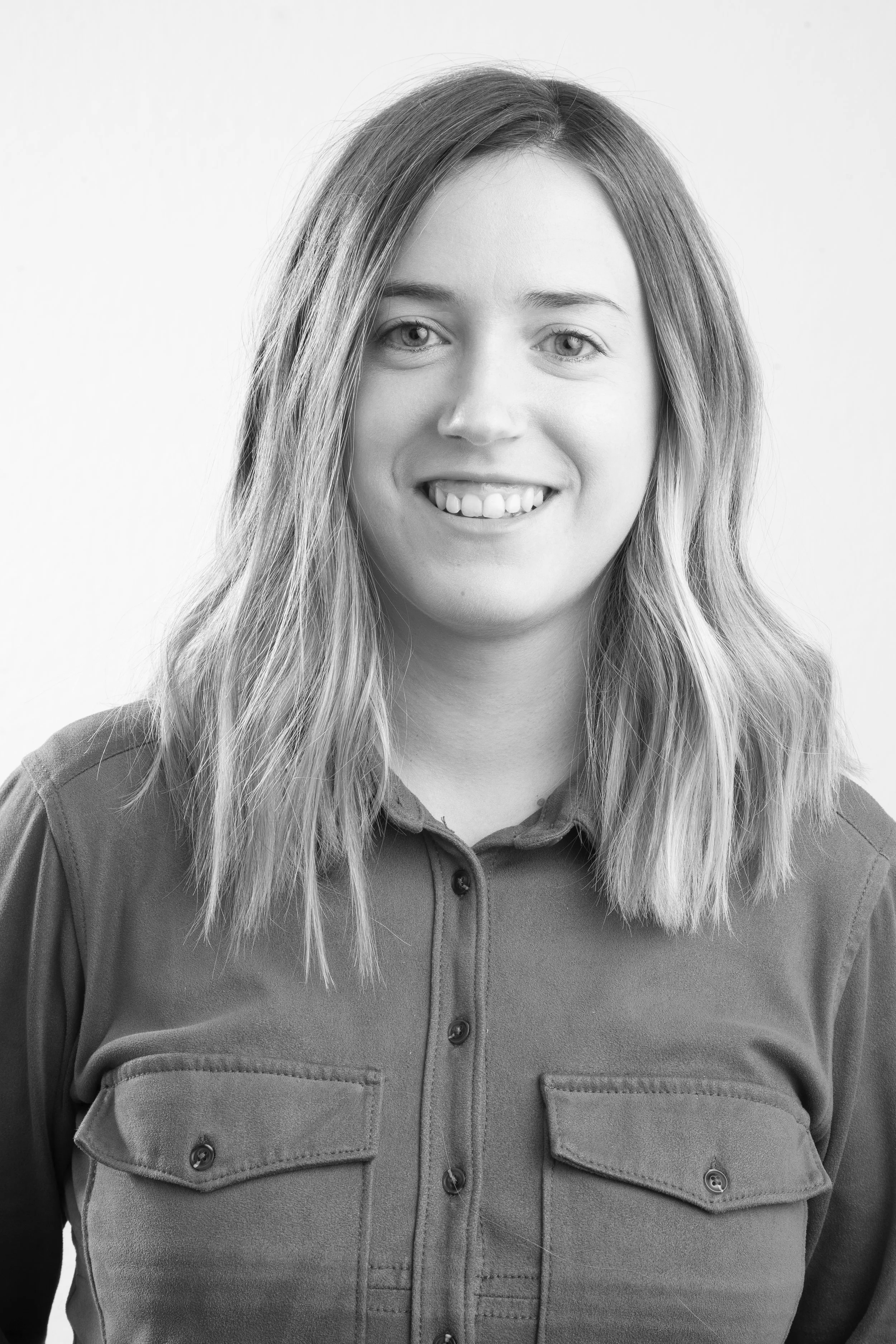 Retrato en blanco y negro de una mujer con cabello rubio y largo, sonriendo, usando una camisa o chaqueta con bolsillos frontales, en un fondo blanco.
