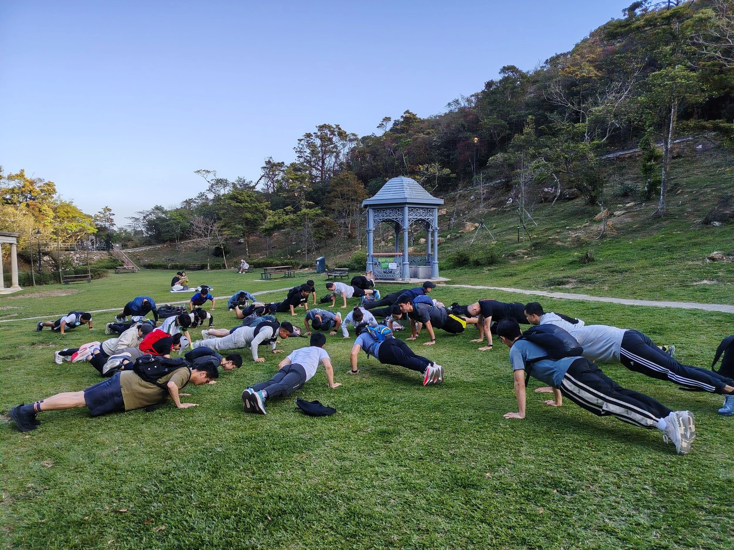 Group of young people doing push-ups on the grass in a park with a small decorative pavilion, trees, and a hillside in the background during the daytime.