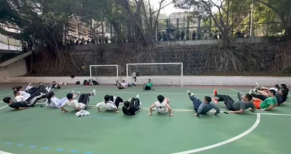 Group of children and teens participating in outdoor physical activity on a sports court with goals, some lying on the ground in a line, under a tree-lined area.