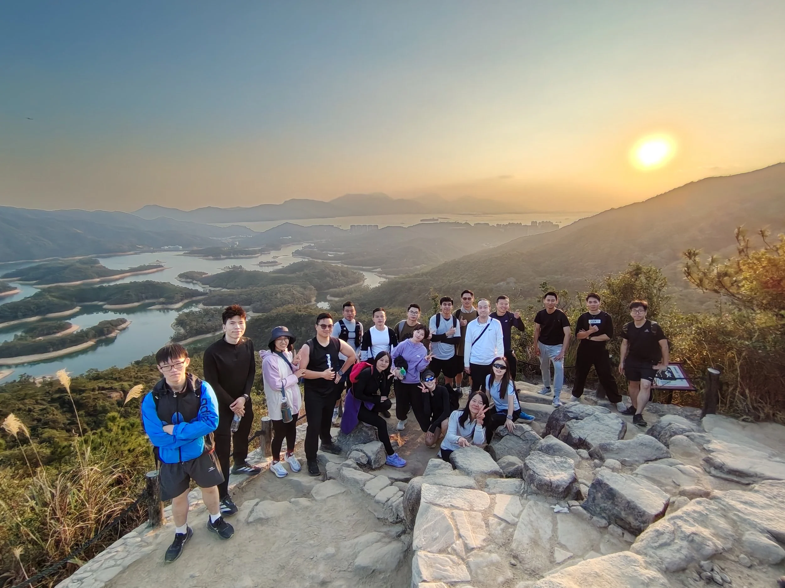 A large group of people standing on rocky terrain at a scenic overlook with mountains, water, and a setting sun in the background.