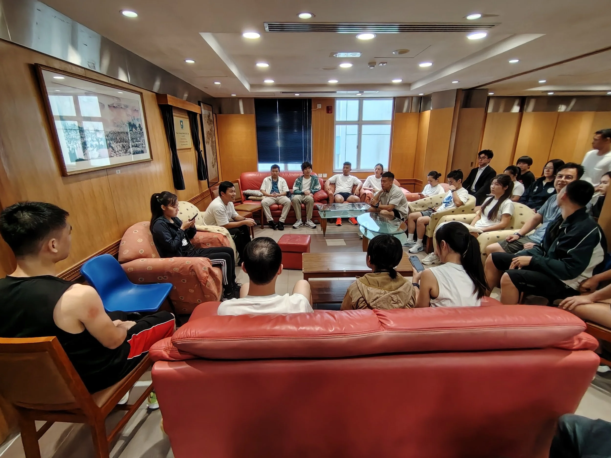 A group of people sitting in a room with wood-paneled walls, some on sofas and chairs, participating in a discussion or meeting, with a woman using her phone in the foreground.