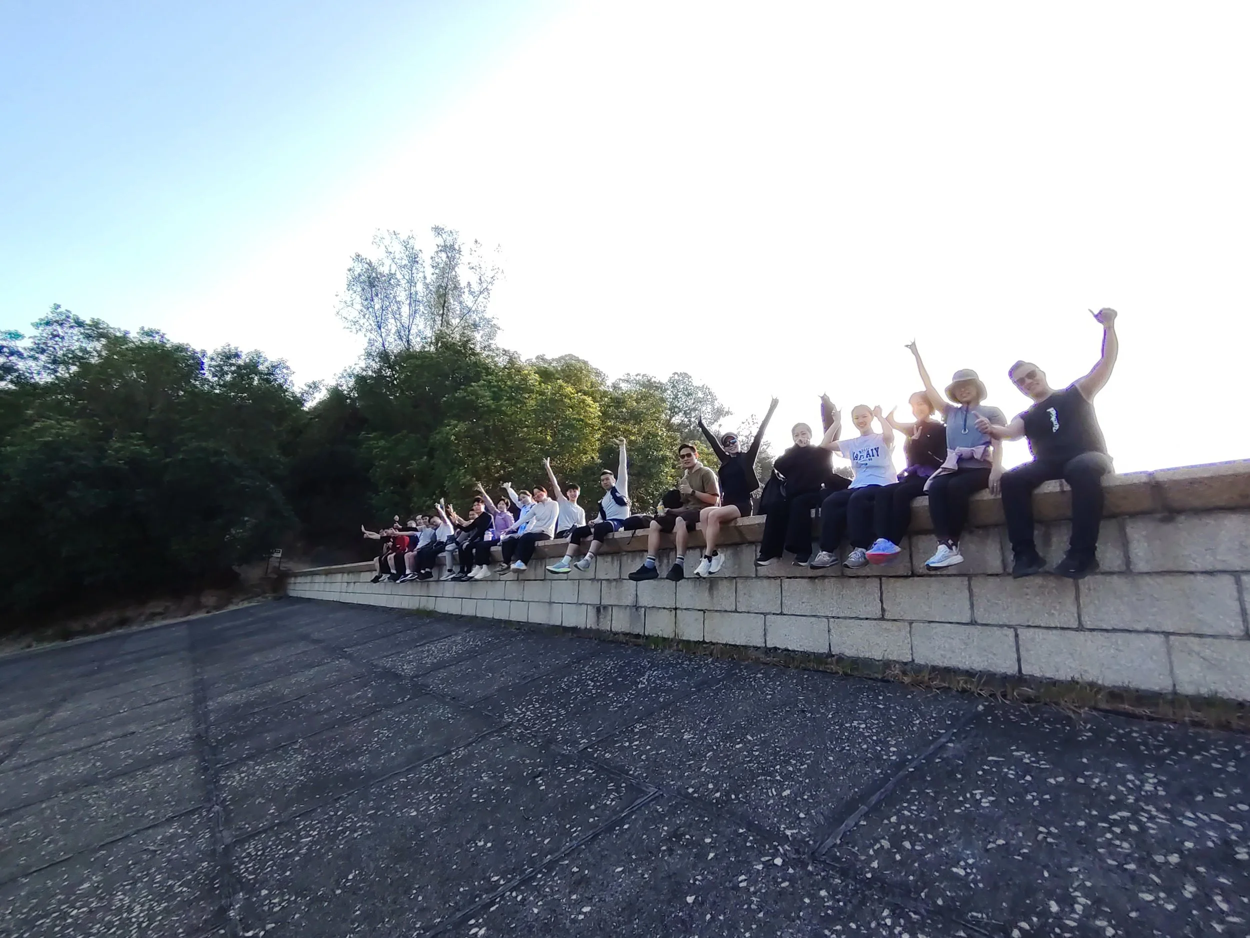 Group of people sitting on a stone ledge in a park, raising their arms and smiling.