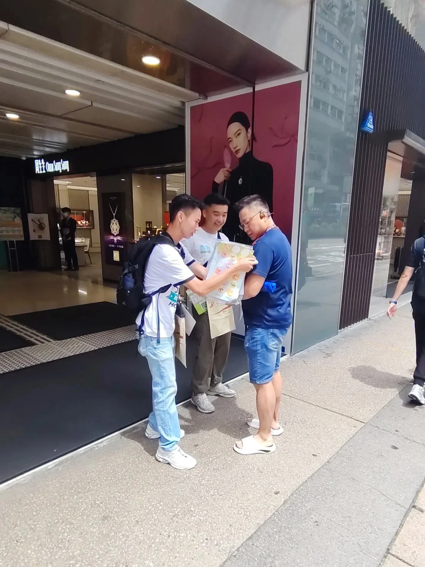 Three young men and a woman standing outside of a building, looking at a colorful piece of paper or a brochure. The building has black and metallic paneling, with a large advertisement poster of a woman in black clothing holding a pink object. One man is wearing a white shirt with a backpack, another in a black shirt, and the third in a blue shirt and shorts with glasses.