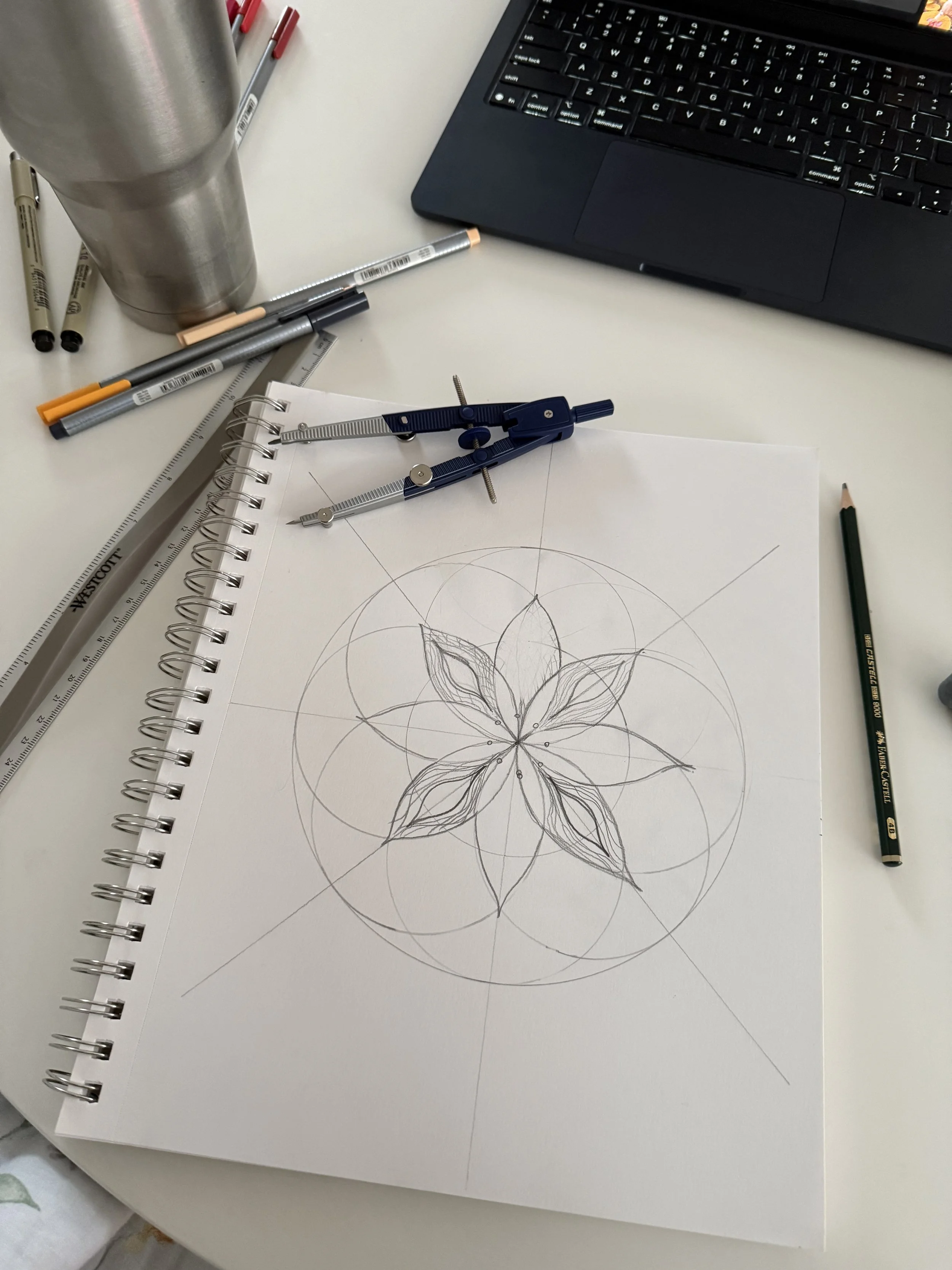 A mandala drawing in progress with pencils and drawing tools beside the sketchbook.