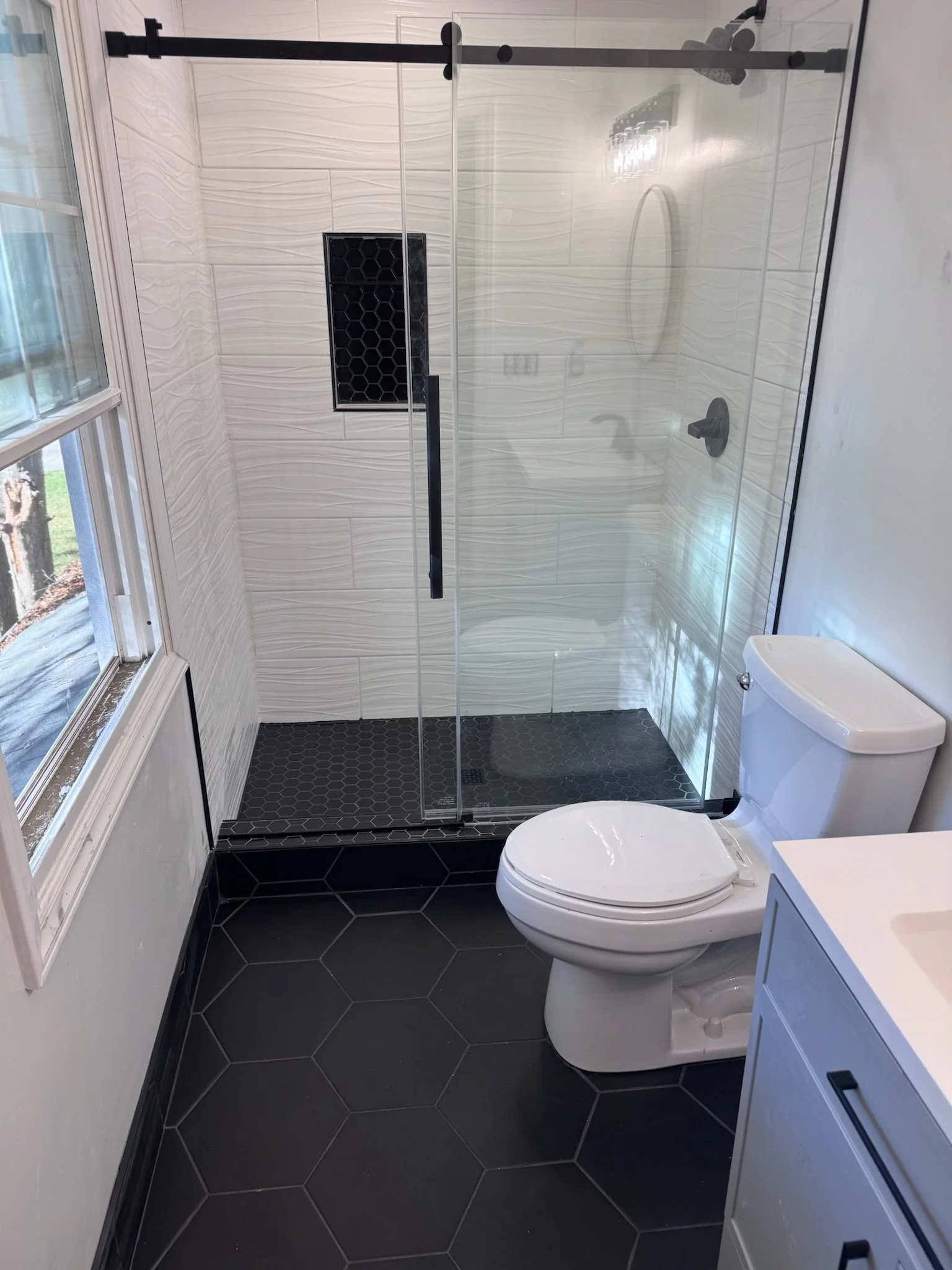 A modern bathroom with a glass shower enclosure, white textured wall tiles, black hexagon floor tiles, a toilet, and a window letting in natural light.