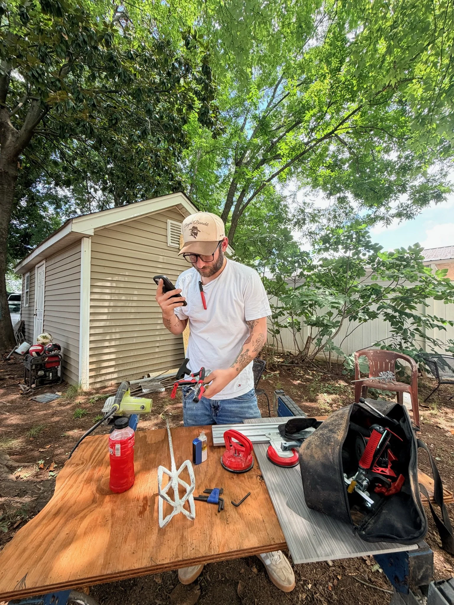 A man with glasses, a beige cap, and tattoos on his arms is working with tools outdoors under a tree, surrounded by equipment and a wooden table with various objects, including a white geometric model and bottled drinks.