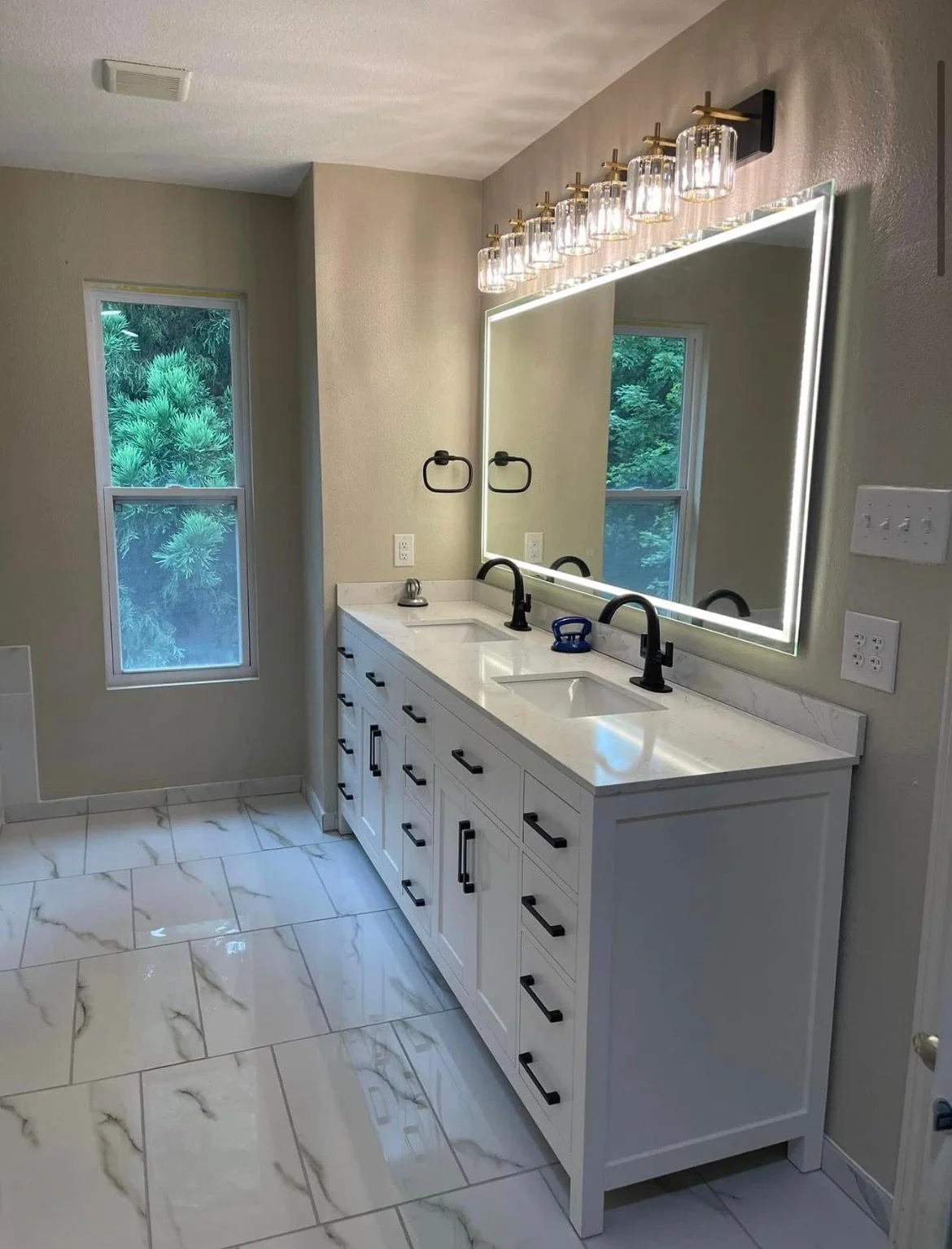 Modern bathroom with double sink vanity, large illuminated mirror, black fixtures, and white marble floor tiles.