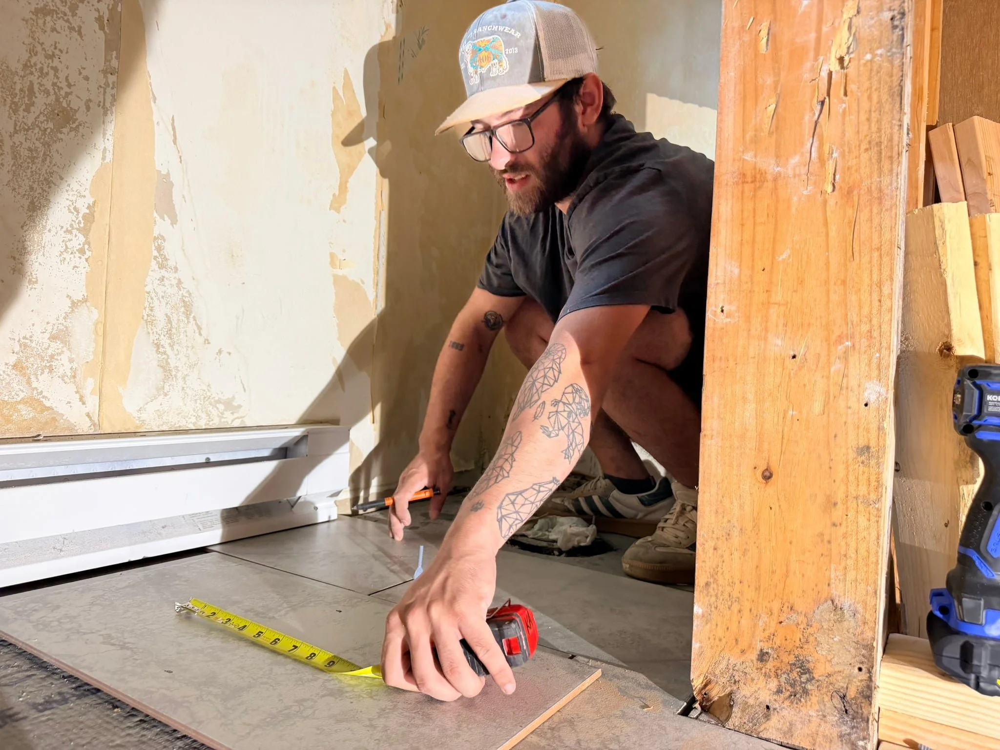 Man wearing glasses and baseball cap crouching on floor, measuring tile with a tape measure, in a room under renovation with peeling paint and exposed wood.
