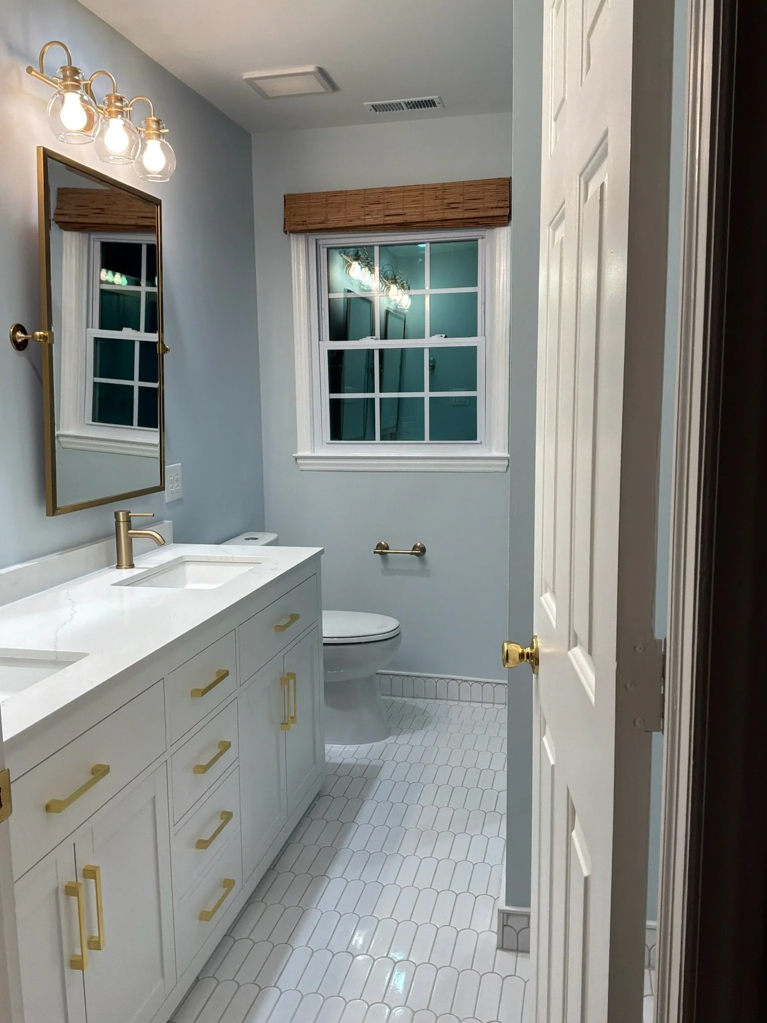 A bathroom features white cabinetry with gold handles, a white countertop with a built-in sink, a gold faucet, a large mirror with a gold frame, a wall-mounted light fixture with four clear glass shades, a window with white trim and a bamboo blinds, and a white hexagonal tiled floor.