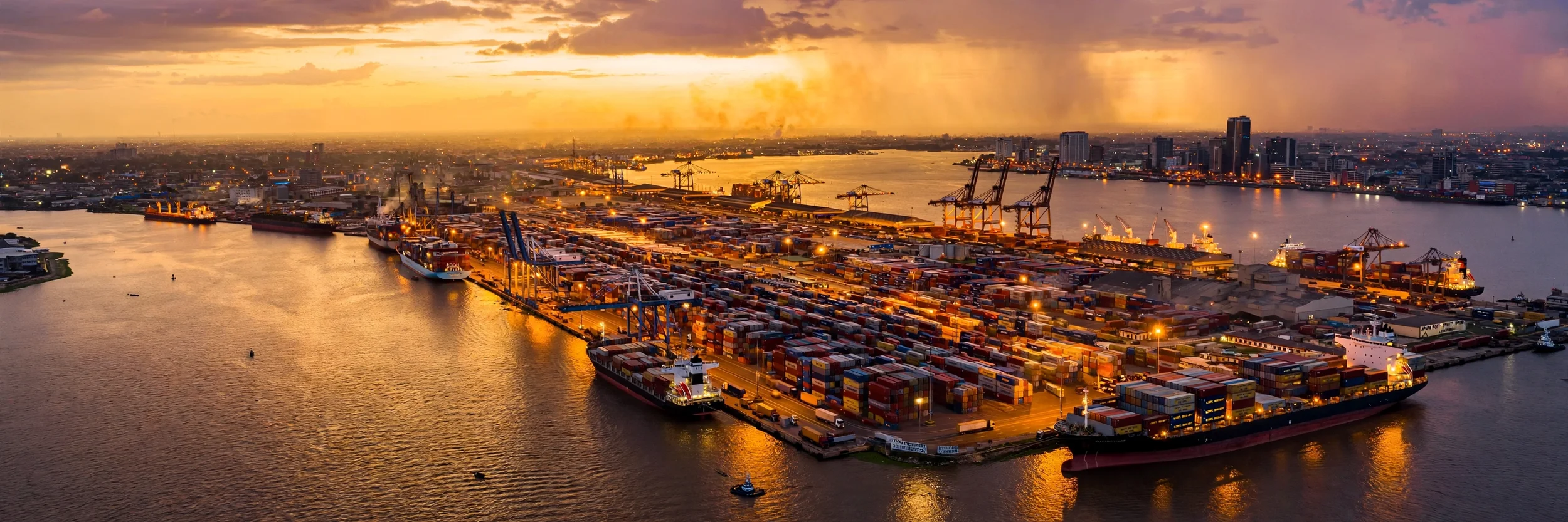 An aerial view of a busy port at sunset with cargo ships docked, cranes, containers, and a city skyline in the background.