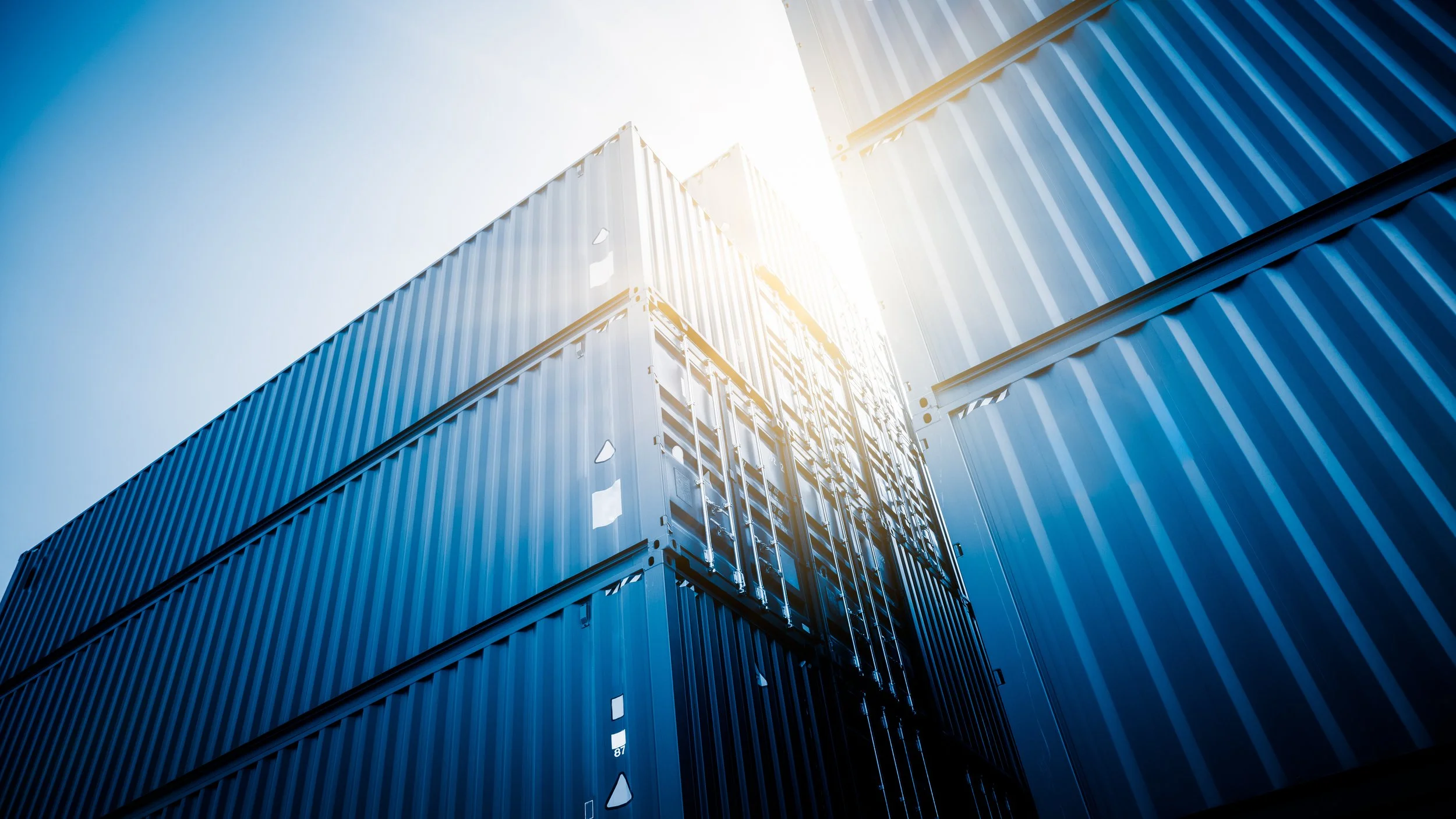 Multiple stacked blue shipping containers viewed from below with sunlight shining from the top right.