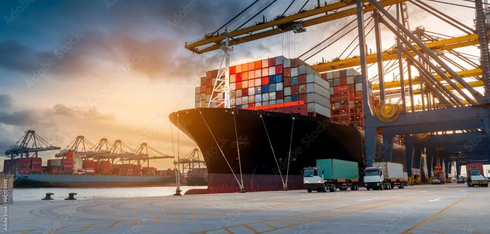 A cargo ship docked at a port with stacks of containers on board, surrounded by cargo trucks and large cranes during sunset.