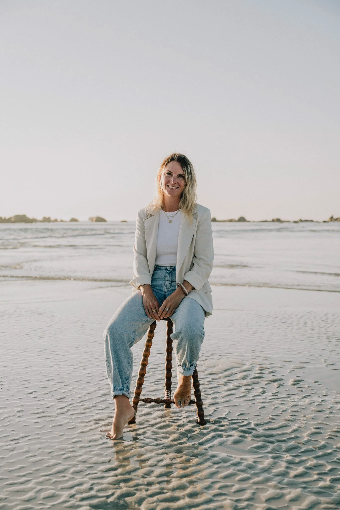 Femme assise sur une chaise en bois au bord de la mer, portant un blazer blanc, un t-shirt blanc et un jean, en équilibre sur une balançoire de bois, sable et mer en arrière-plan, coucher de soleil