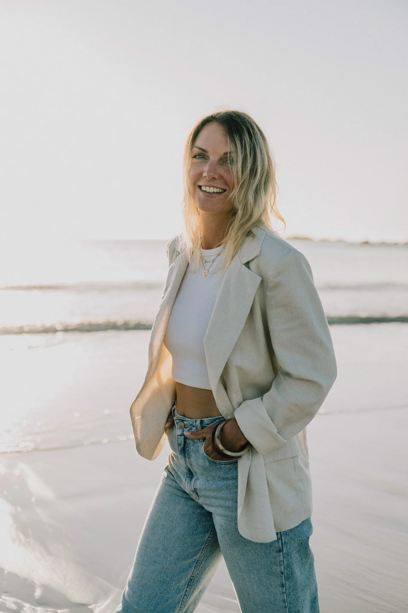 Femme souriante portant une veste beige, un t-shirt blanc et un jean, debout sur la plage avec la mer en arrière-plan au coucher du soleil.