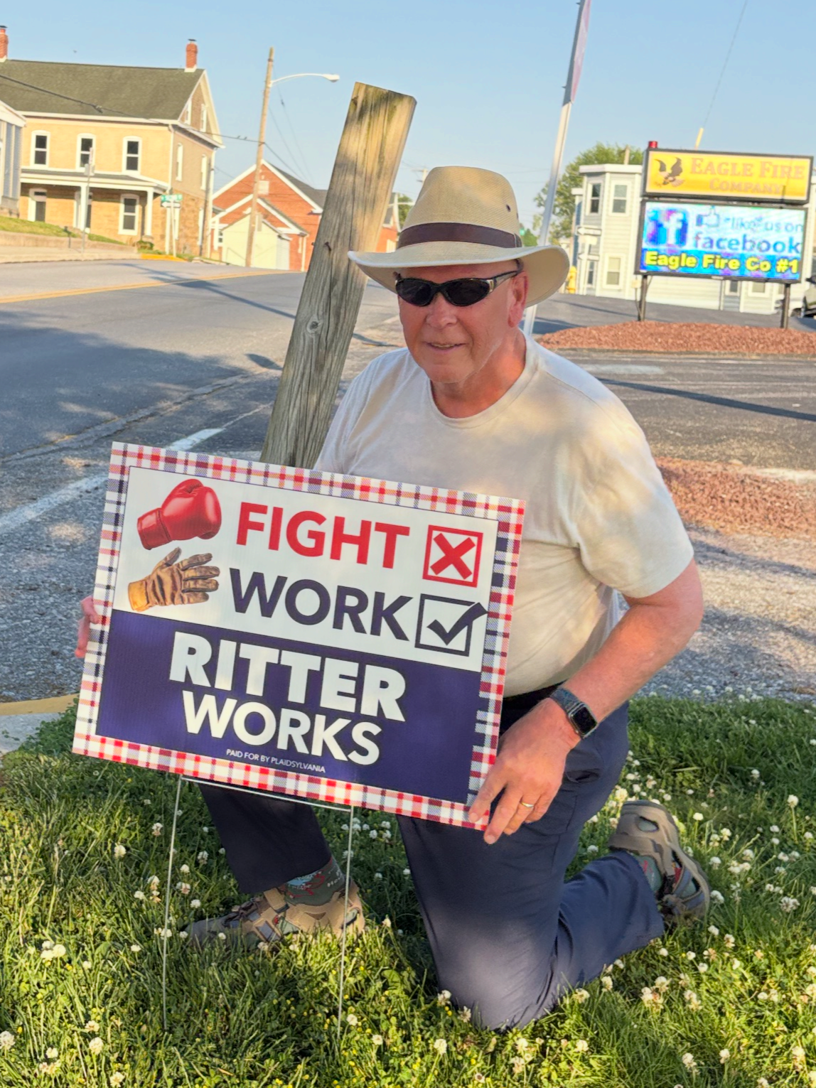 Edward Ritter kneeling on grass with small white flowers, holding a campaign sign. Edward is wearing a beige hat, sunglasses, a aplaid shirt, and gray pants. In the background, there is a street, houses, and a sign for Eagle Fire Company.