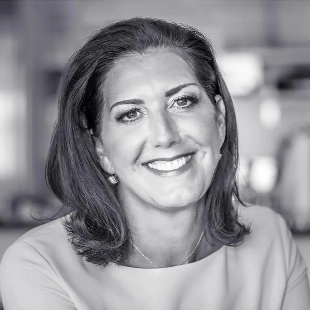 Black and white portrait of a smiling woman with shoulder-length dark hair, wearing earrings, a necklace, and a light-colored top, in an indoor setting.