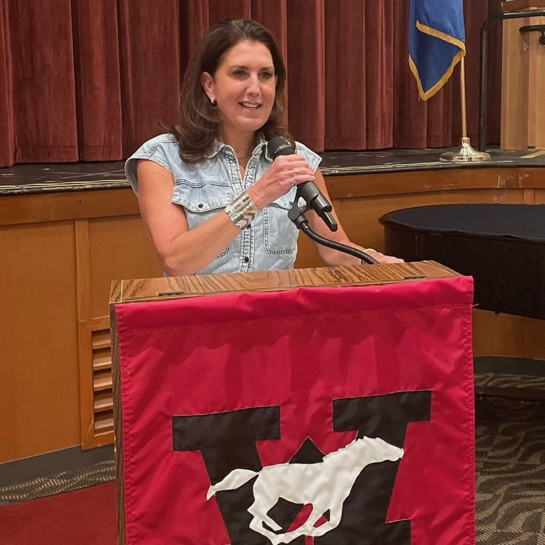 A woman is speaking at a podium with a red cloth and a black logo of a horse's head, in a room with wooden walls, a stage, and a blue flag in the background.