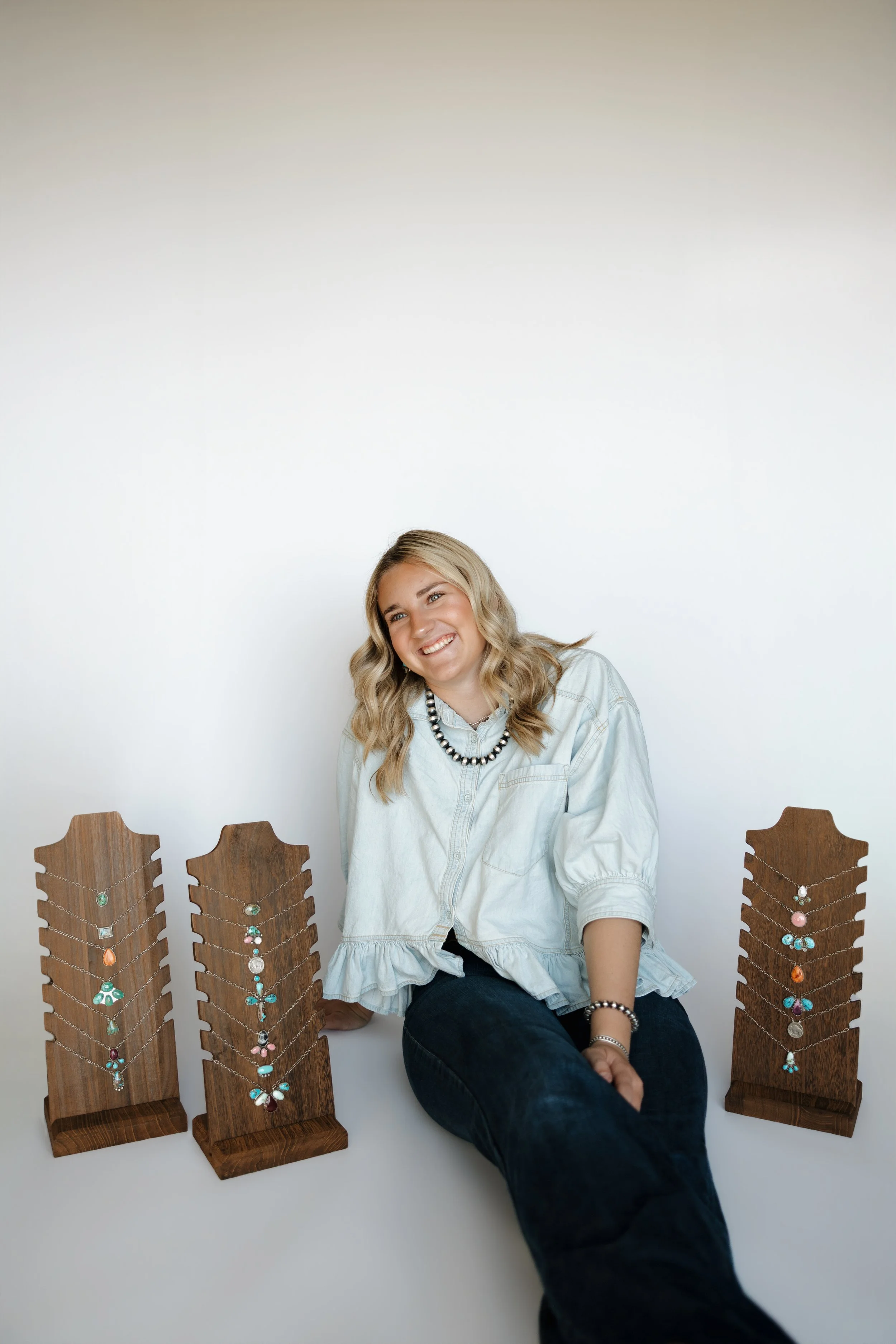 A woman sitting in front of a white wall with wooden jewelry displays on either side, featuring colorful necklaces.