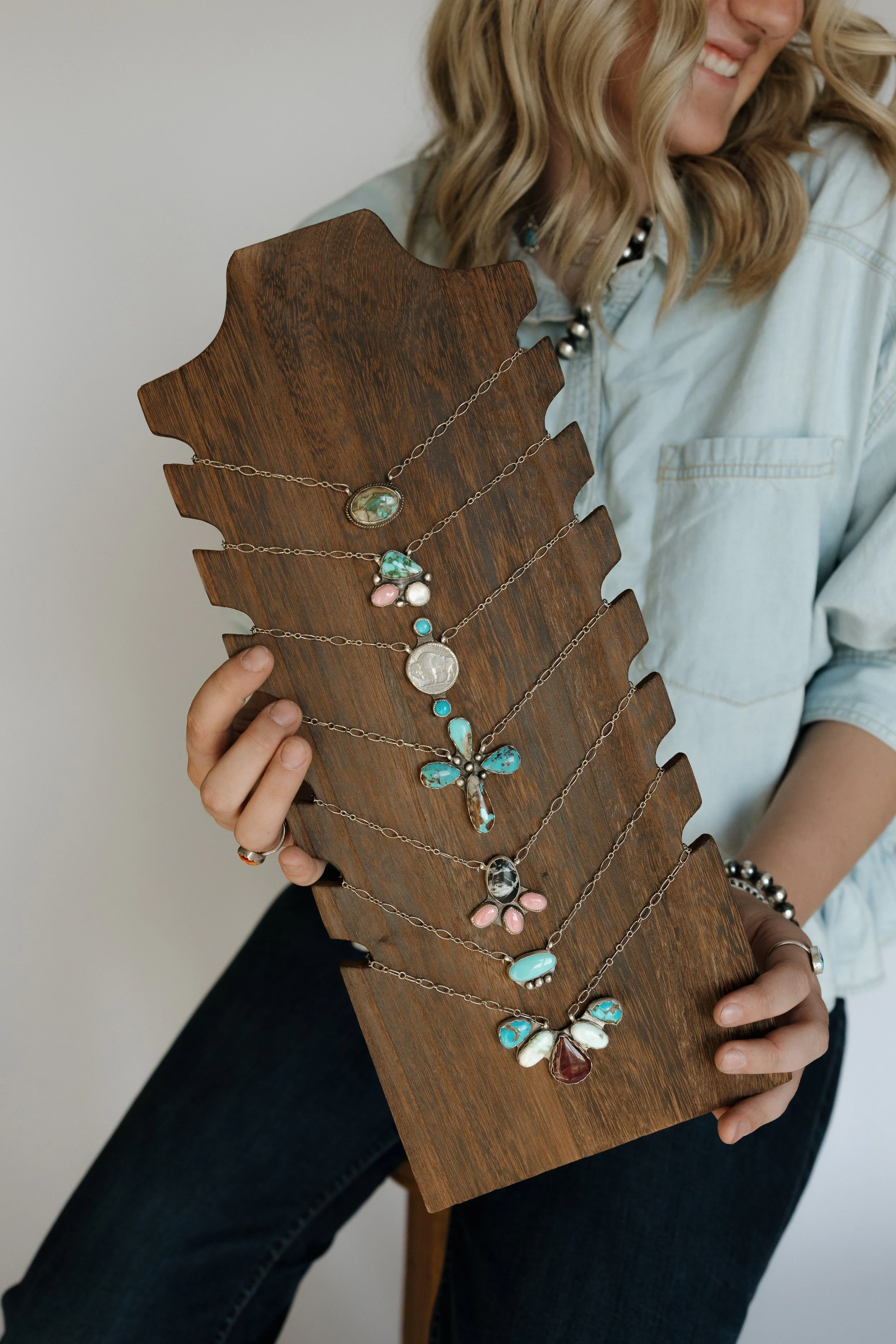 Person with wavy blonde hair wearing a light denim shirt and black pants, holding a wooden display with several layered necklaces featuring various colorful pendant designs.