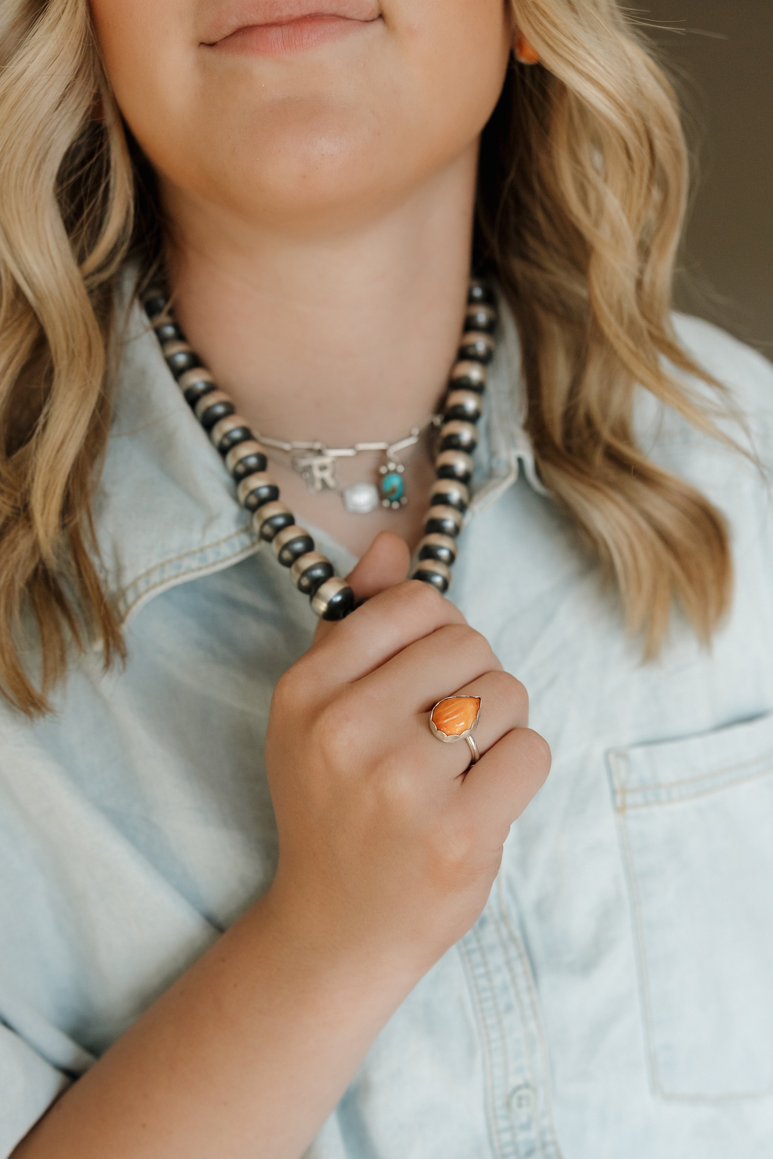 Close-up of a woman wearing a pearl necklace and a silver ring with a large orange gemstone, dressed in a light denim shirt.