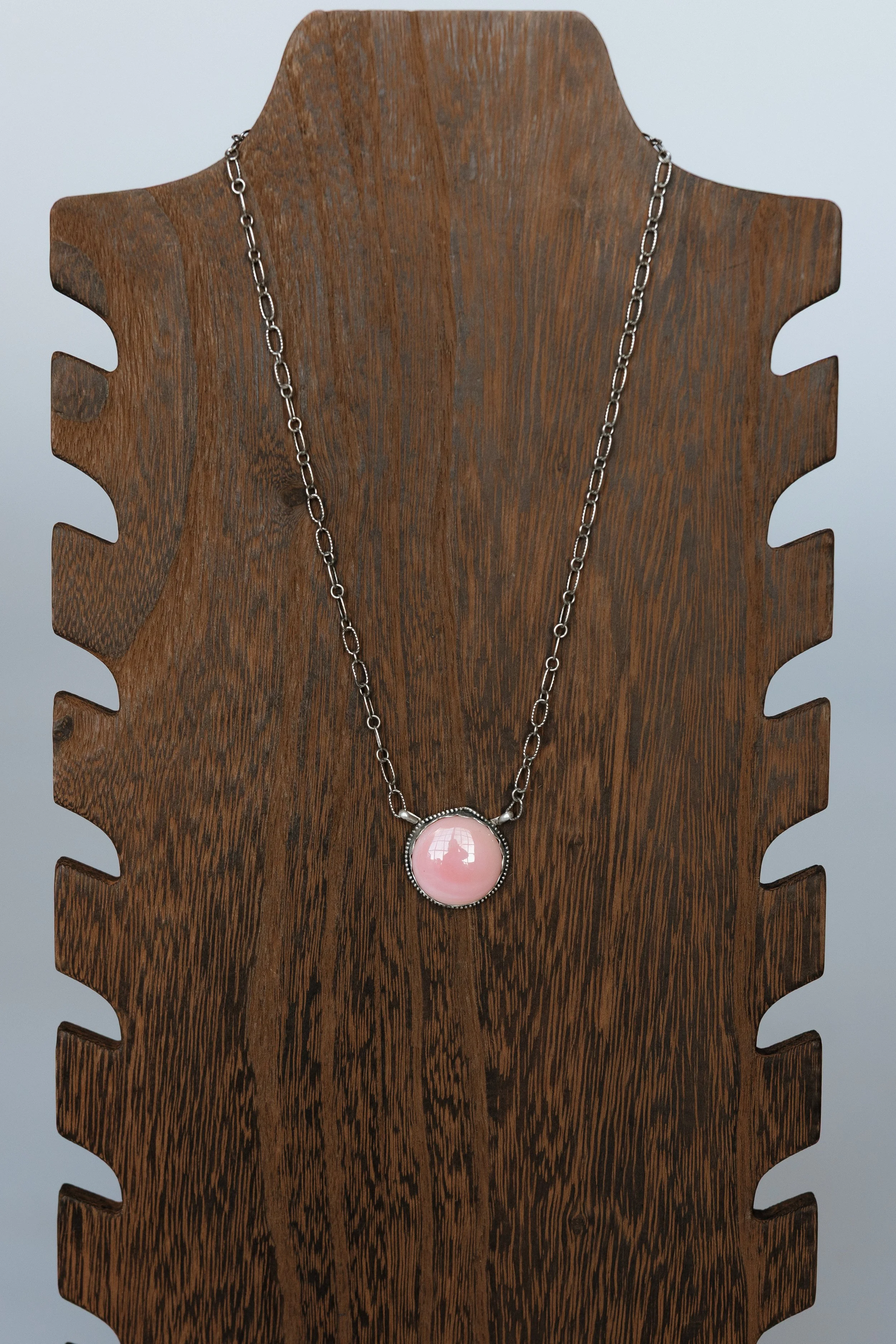A silver necklace with a pink oval pendant hanging on a wooden display stand.