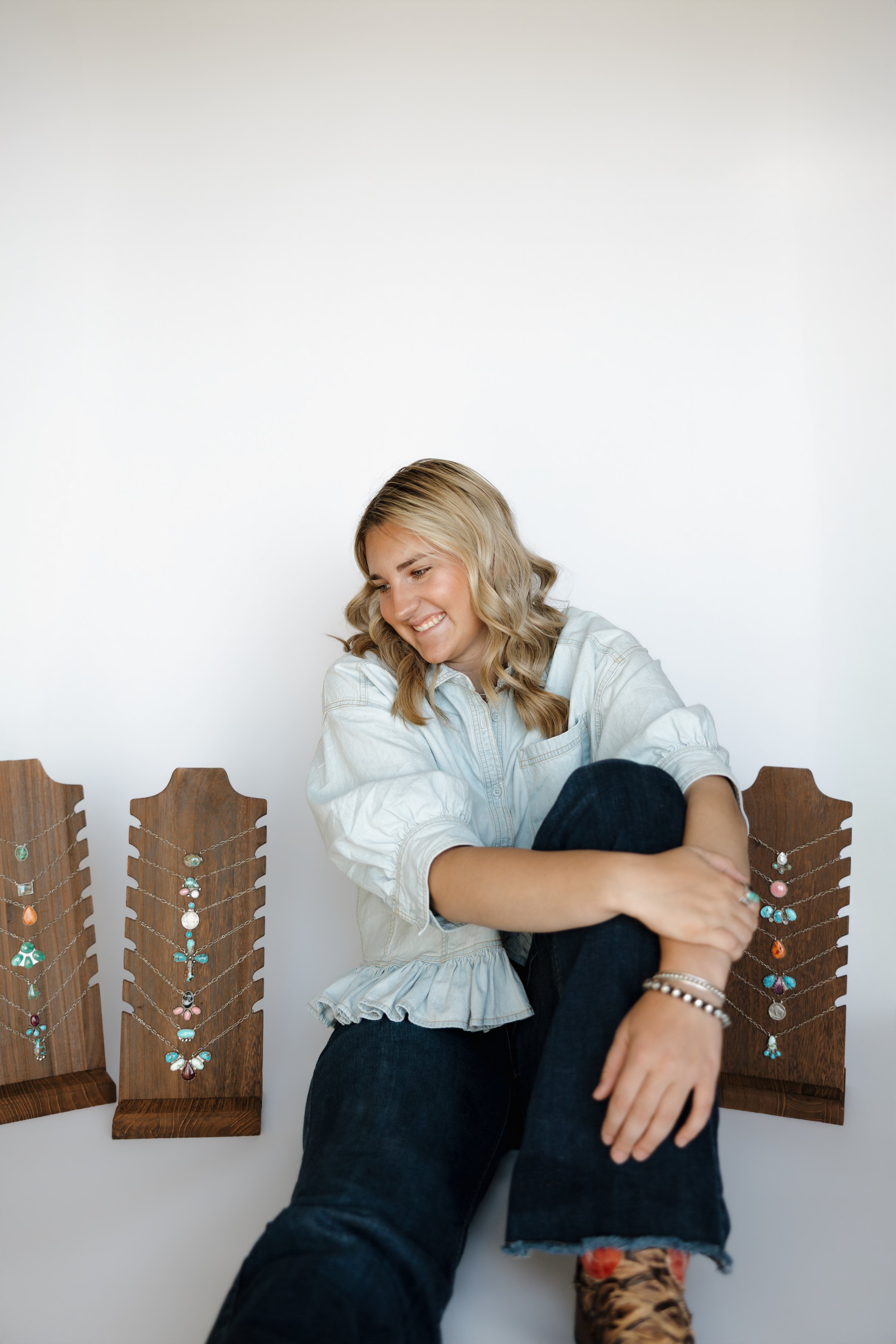 A woman sitting on a wooden chair with a jewelry display, smiling and looking down, against a plain white wall.