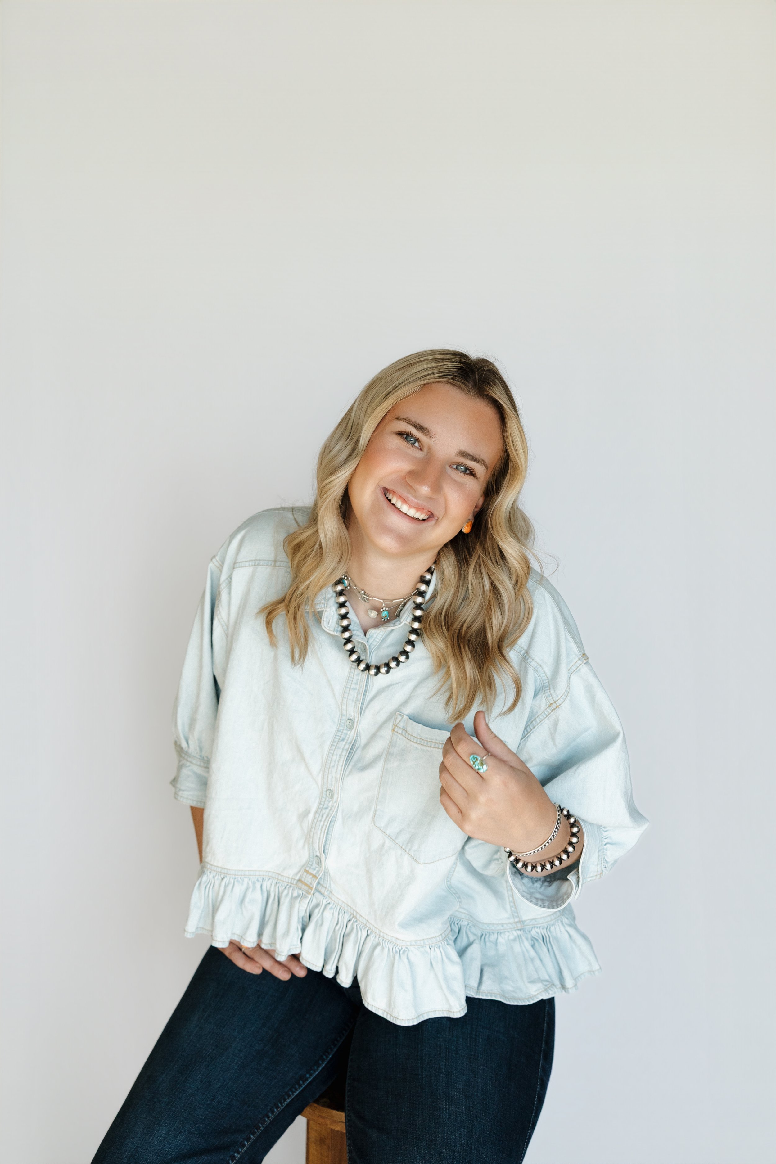 A young woman with blonde wavy hair smiling, wearing a light-colored denim shirt, black jeans, layered necklaces, earrings, a ring, and bracelets, sitting on a wooden stool against a plain white background.