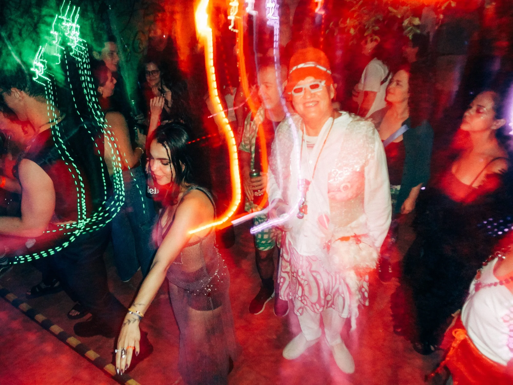 Mulher com vestido brilhante dançando, outras pessoas ao redor em movimento, iluminação de luzes coloridas, ambiente festivo ou de celebração noturna.