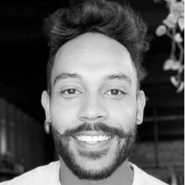 Retrato de um homem sorridente com barba e cabelo cacheado, em fundo escuro.