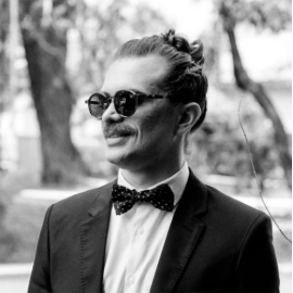 Homem com cabelo preso, usando óculos escuros, terno e gravata borboleta, sorrindo ao ar livre