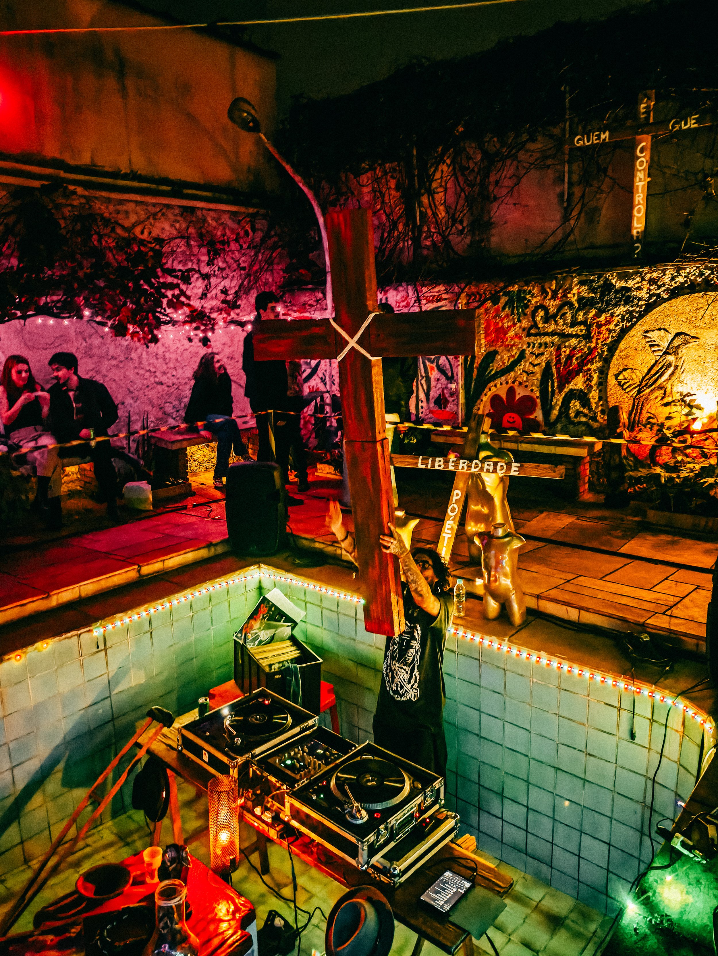 Cena de uma festa com DJ tocando músicas, pessoas sentadas e conversando, decoração com luzes coloridas, uma cruz de madeira ao centro e uma parede com grafite e pintura artística.