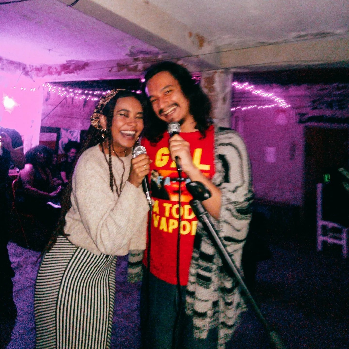 Dois indivíduos sorrindo, segurando microfones, em ambiente com luzes coloridas e decoração festiva.