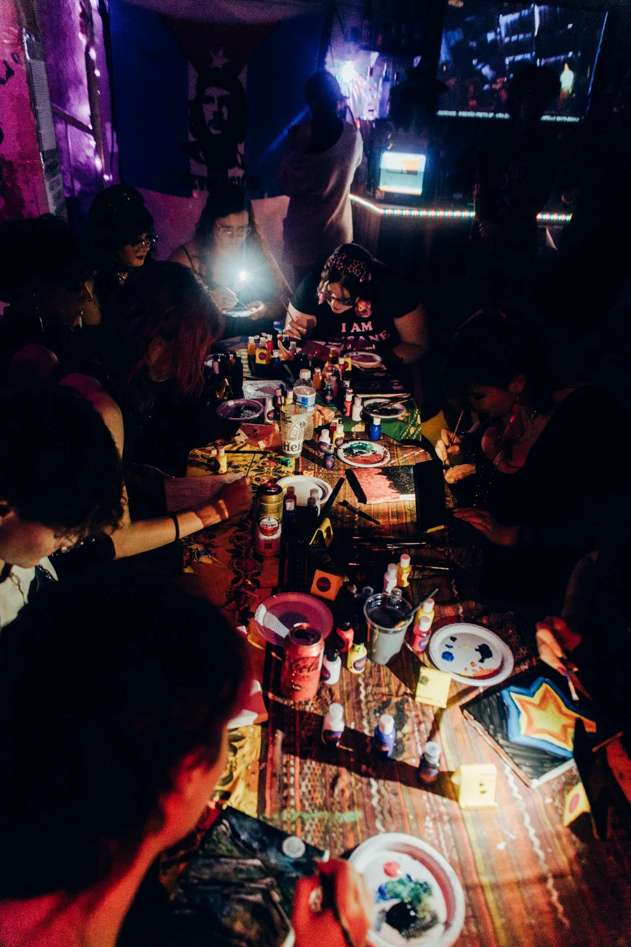 Reunião de várias pessoas em uma mesa, realizando pintura ou artesanato, com vários recipientes de tinta, pincéis e materiais variados, em ambiente com iluminação baixa.