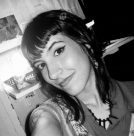 Retrato de uma jovem com cabelo curto e escuro, usando uma ceia com detalhes em branco, sorrindo para a câmera, em uma ambiente interno.