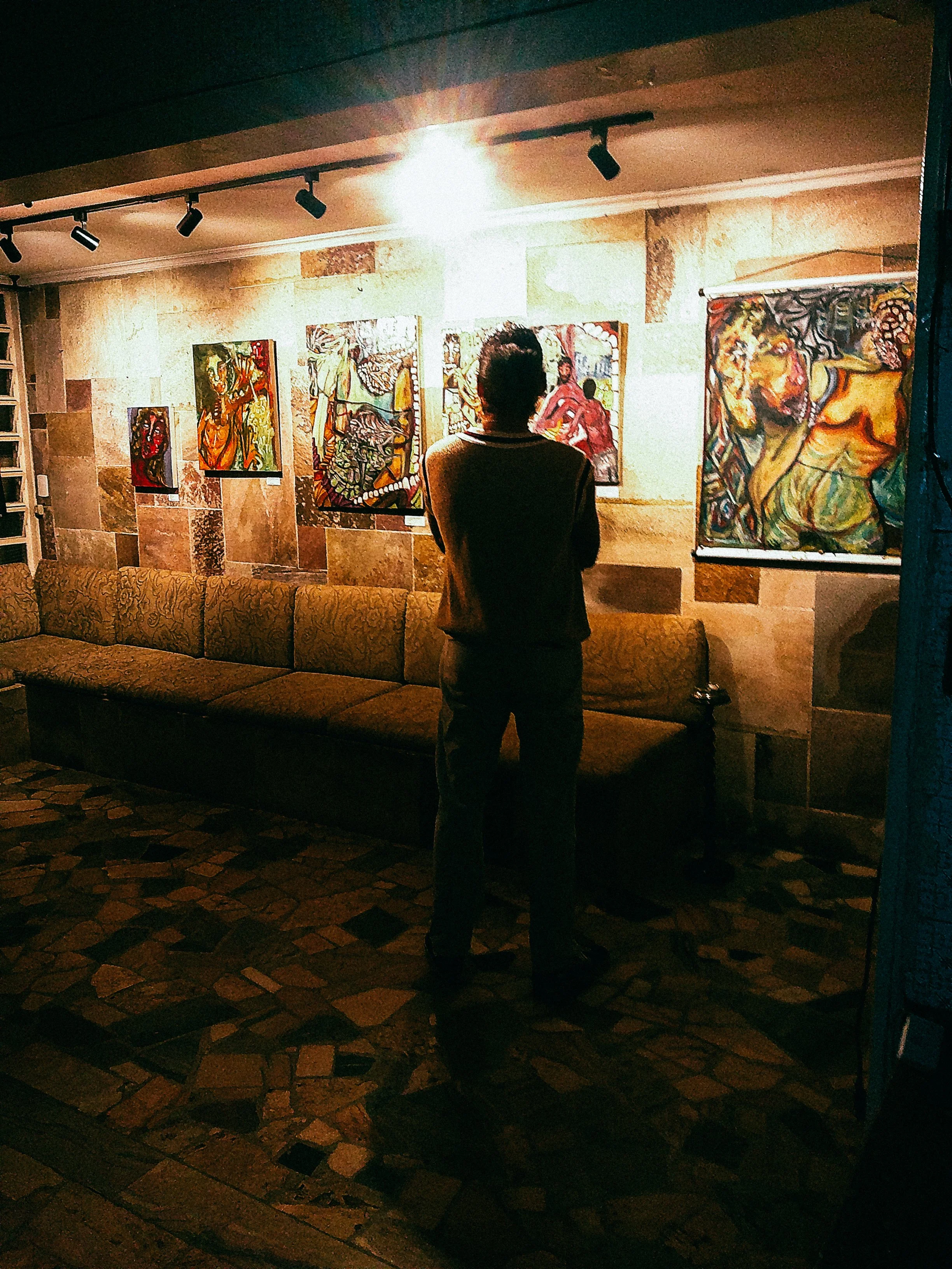 Pessoa assistindo uma exposição de quadros em um salão com iluminação direcionada, paredes de pedra e uma cadeira longa.
