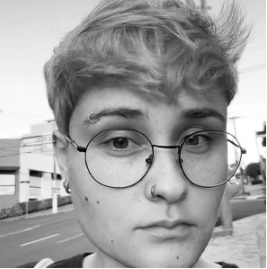 Retrato de uma jovem com cabelo castanho claro, usando óculos de armação redonda, com expressão séria, em ambiente urbano.