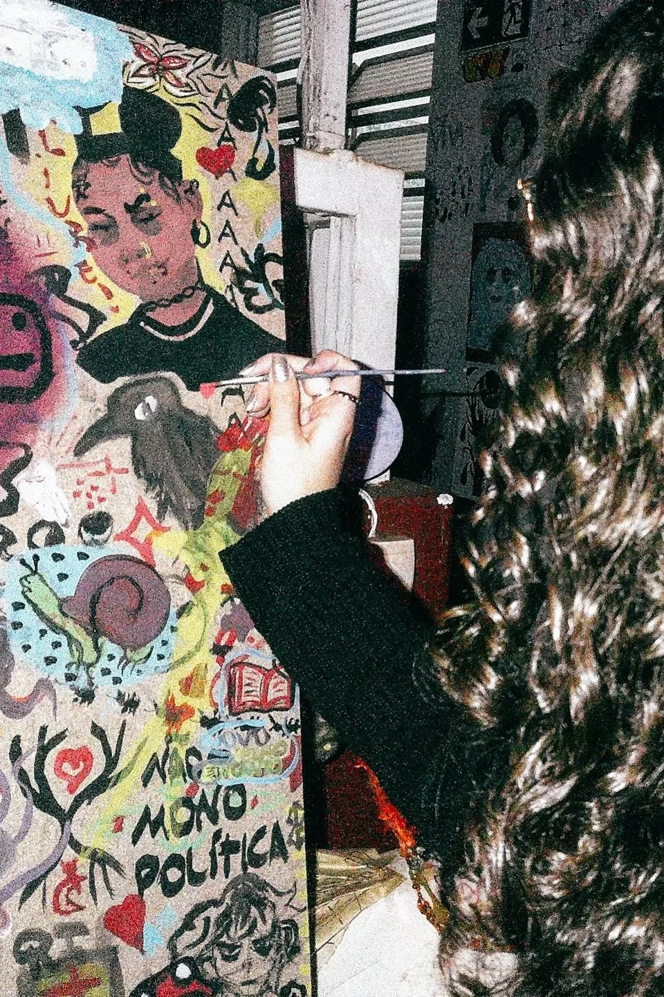 Pessoa pintando uma arte de rua com cores vibrantes que retrata uma mulher com cabelo escuro e curvo, além de letras e desenhos diversos ao redor, enquanto uma pessoa com cabelo cacheado observa.