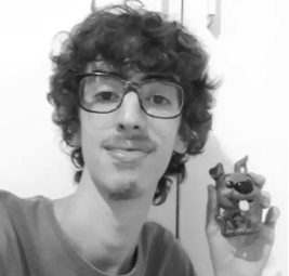 Jovem com cabelo cacheado, óculos e barba, segurando uma figura de brinquedo de um cão, em um ambiente interno