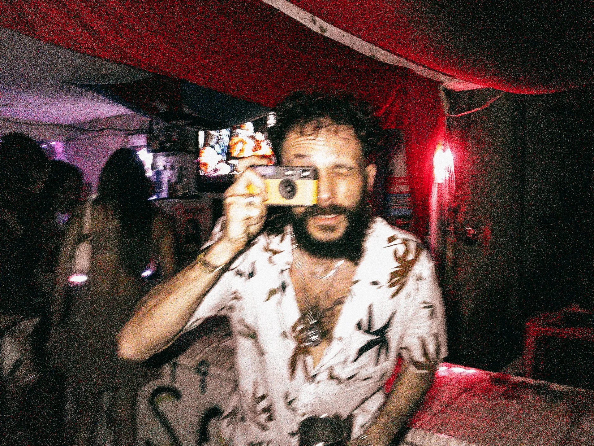 Homem com barba, cabelo cacheado, usando uma camisa clara com estampa de letras, segurando uma câmera vintage em um ambiente com iluminação vermelha, ao lado de uma mulher de cabelo escuro