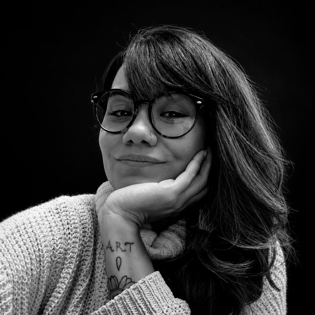 Retrato em preto e branco de uma mulher sorrindo, com óculos de armação grossa, cabelo comprido e uma tatuagem no braço, com fundo preto. Ela está apoiando a cabeça na mão com expressão serena.
