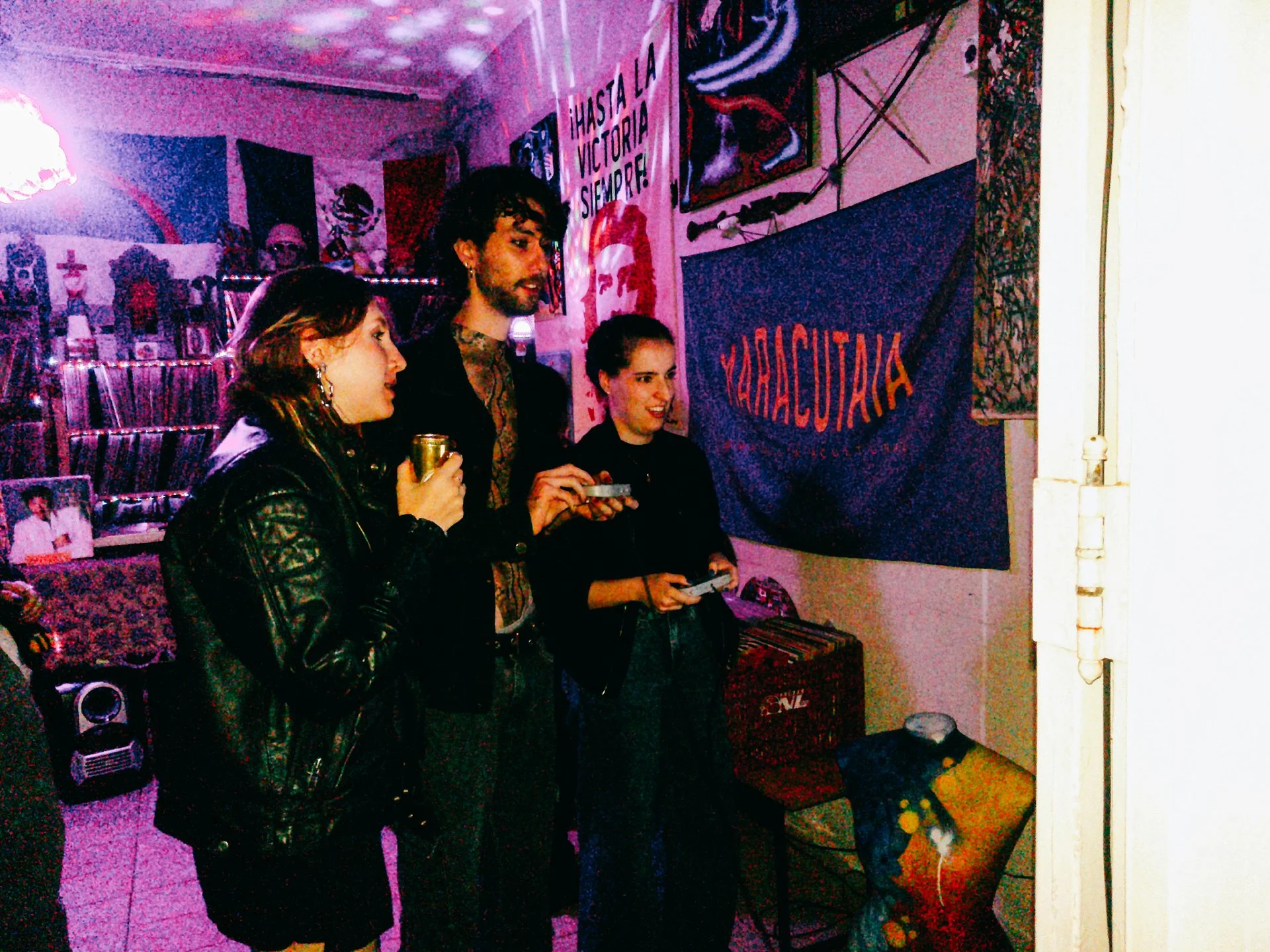 Três pessoas assistindo a algo em um ambiente decorado com bandeiras e pôsteres, incluindo uma bandeira do partido MMA e uma parede decorada com motivos mexicanos.