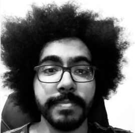 Pessoa com cabelo afro, usando óculos e barba, olhando para a câmera em uma foto em preto e branco.